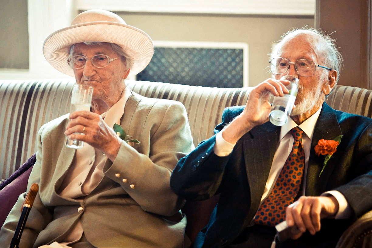 Two elderly wedding guests sipping drinks together on a striped sofa