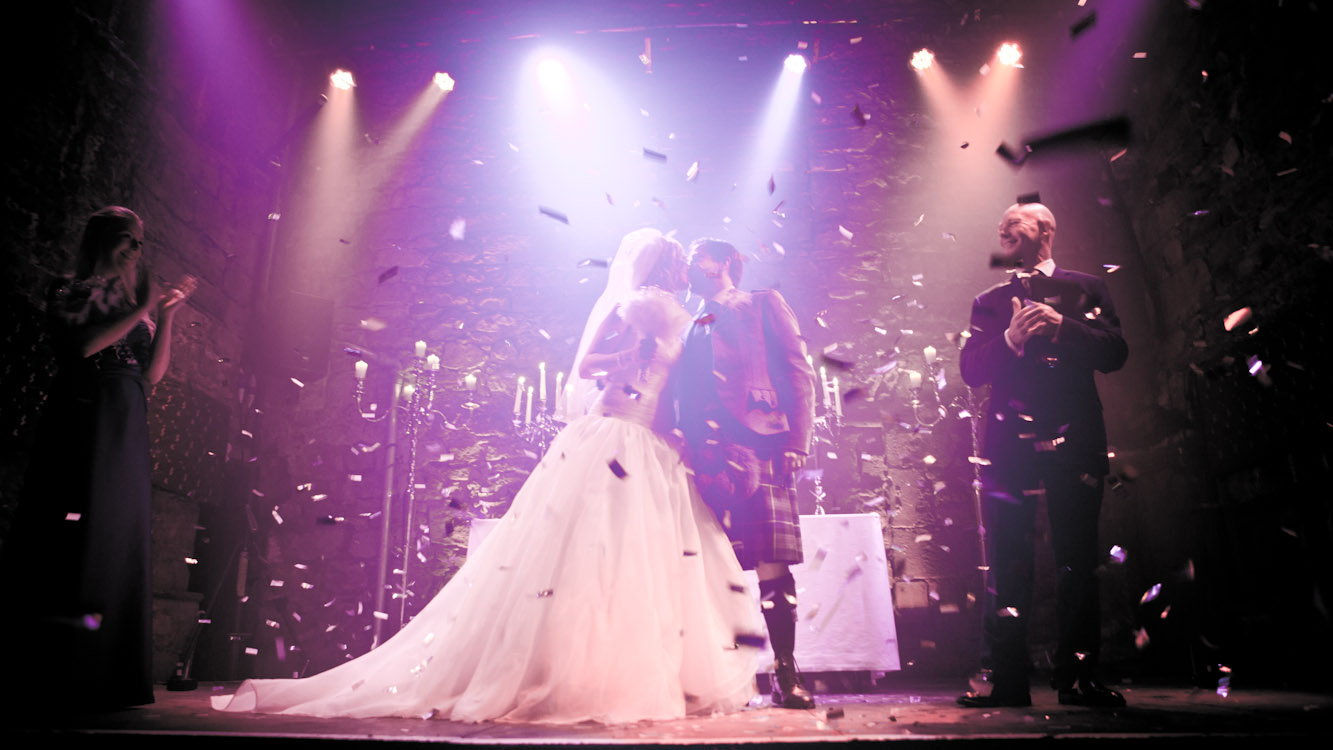 Bride and groom in a kilt kissing under confetti and stage lights at their reception