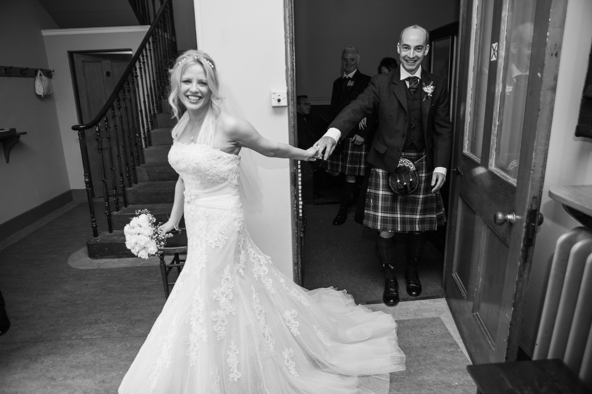 Black and white photograph of a bride leading her kilted groom by the hand through a doorway