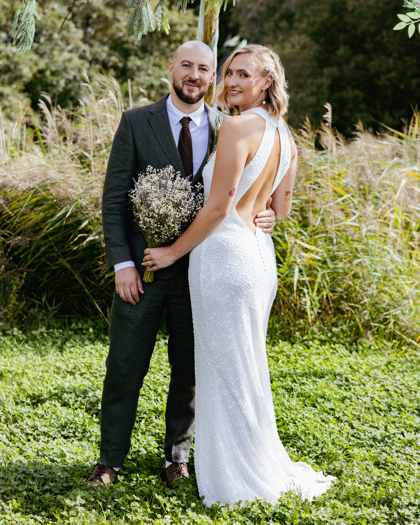 Bride in a beaded gown looking over her shoulder beside her groom holding a dried lavender bouquet