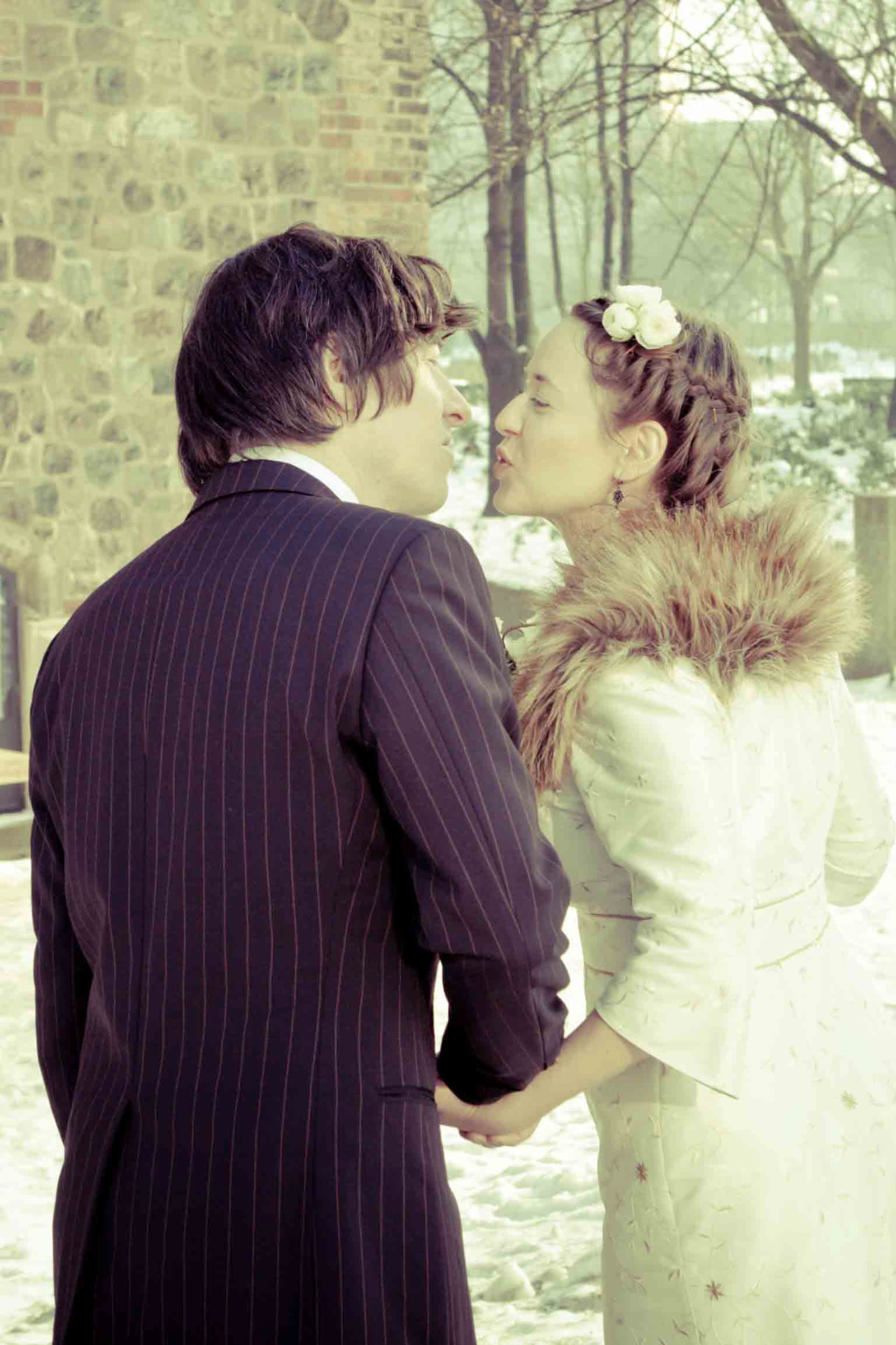 Bride in a fur collar leaning in to kiss her groom in the snow outside a stone church