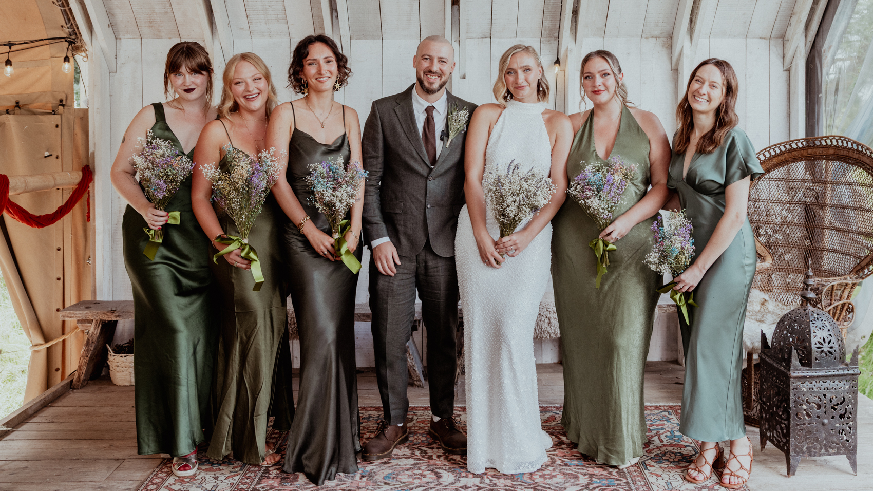 Bride, groom and bridal party in green silk dresses inside a rustic wooden wedding venue
