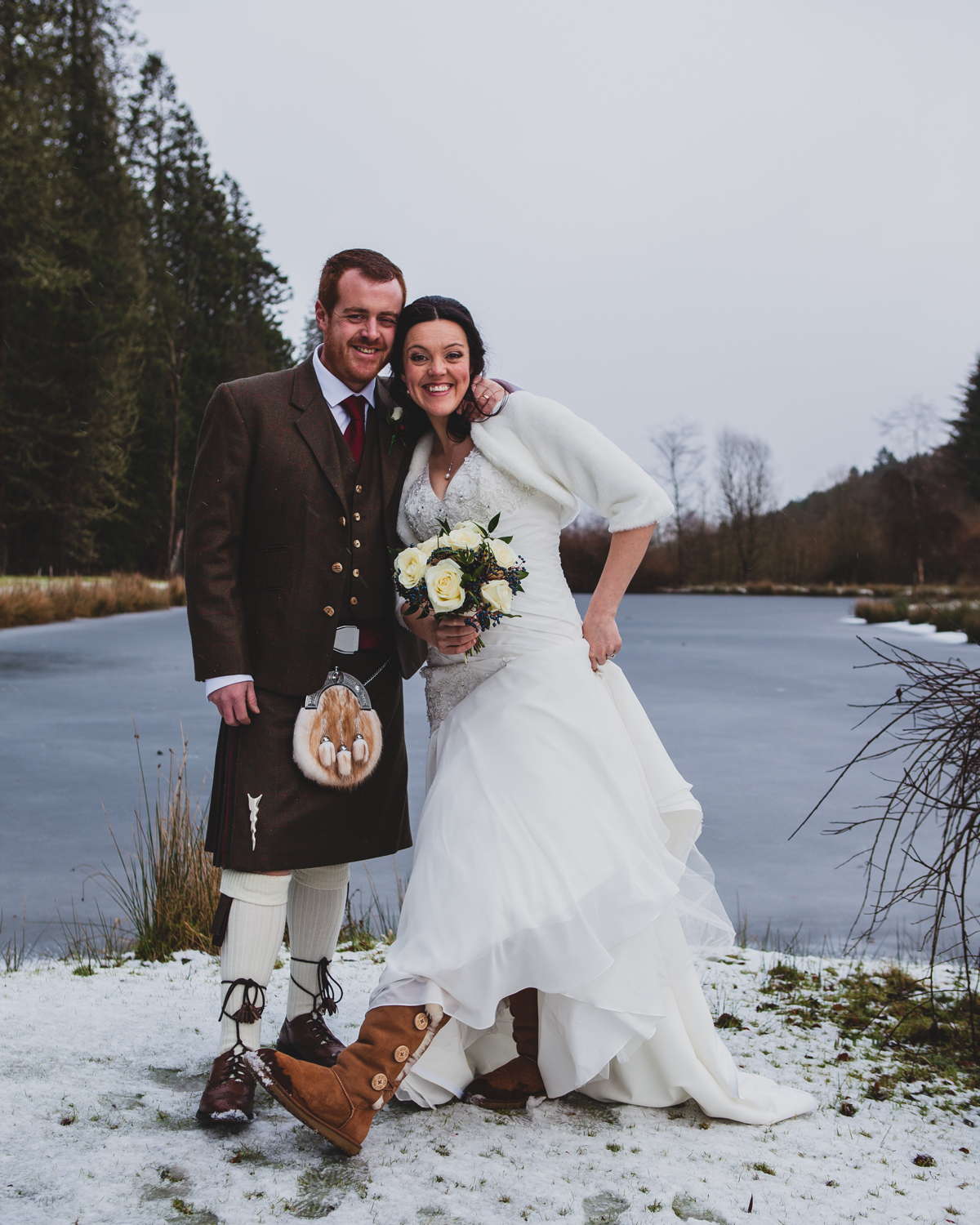 Bride in a fur shrug and groom in a tweed kilt smiling beside a frozen loch in the snow
